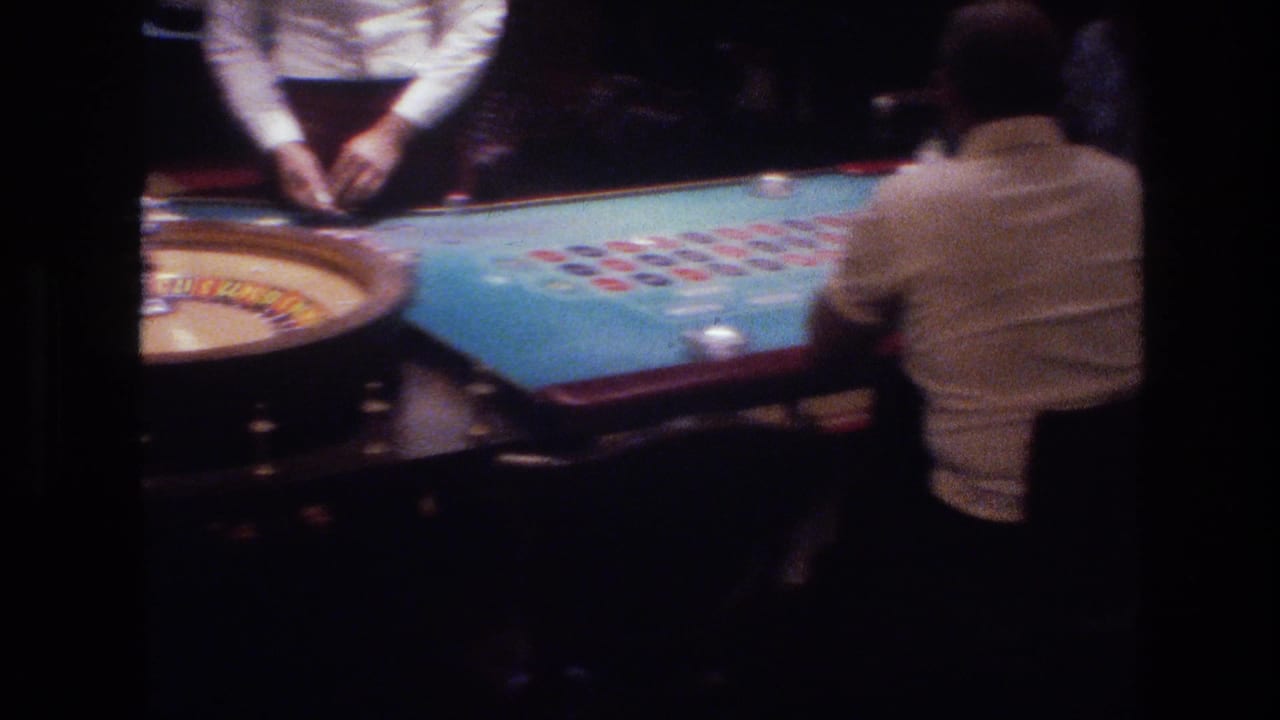 Super 8 shot from behind a patron at a Las Vegas roulette table circa 1977, showing blue felt betting layout, colored chips, and a dealer cropped at chest height.