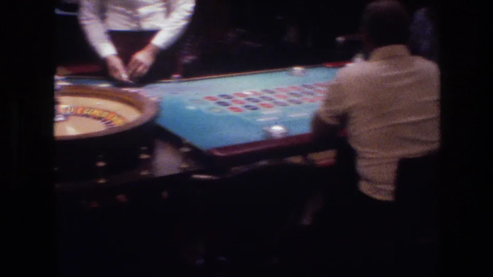 Super 8 shot from behind a patron at a Las Vegas roulette table circa 1977, showing blue felt betting layout, colored chips, and a dealer cropped at chest height.