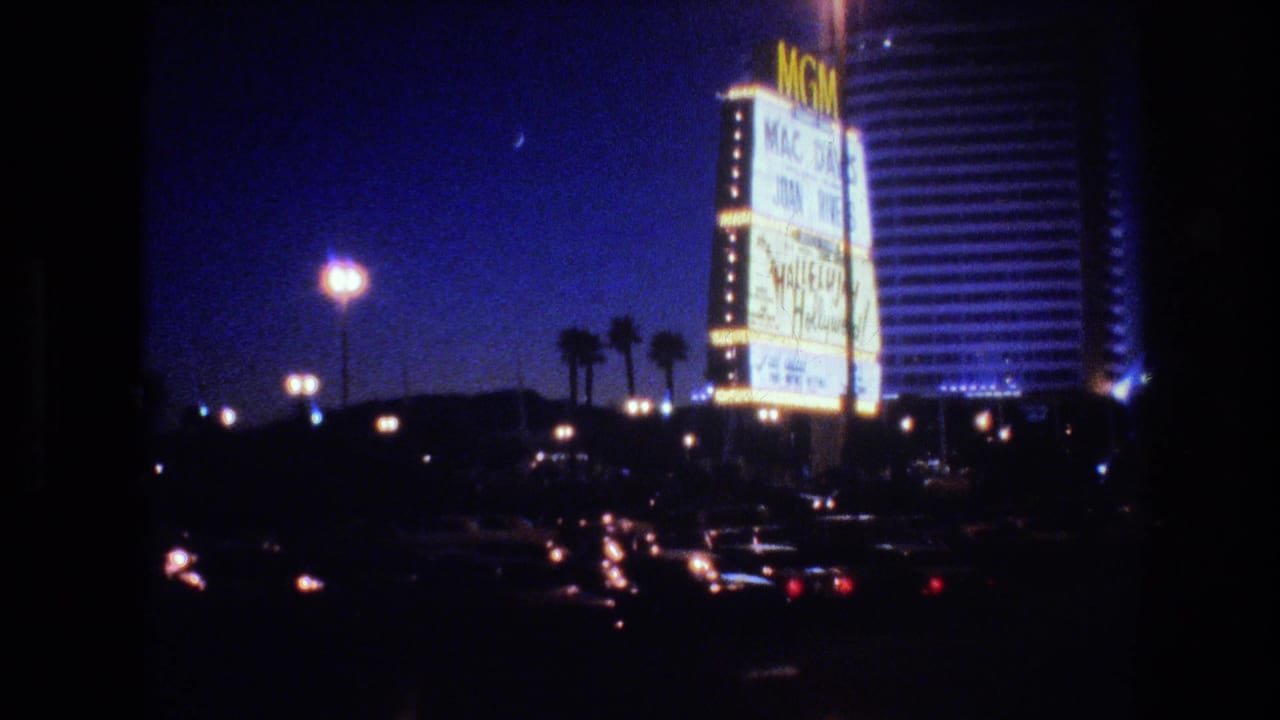 Wider 8mm shot of the MGM Grand hotel on the Las Vegas Strip at night in 1977, with passing cars in the foreground, palm trees, and street lamps visible.