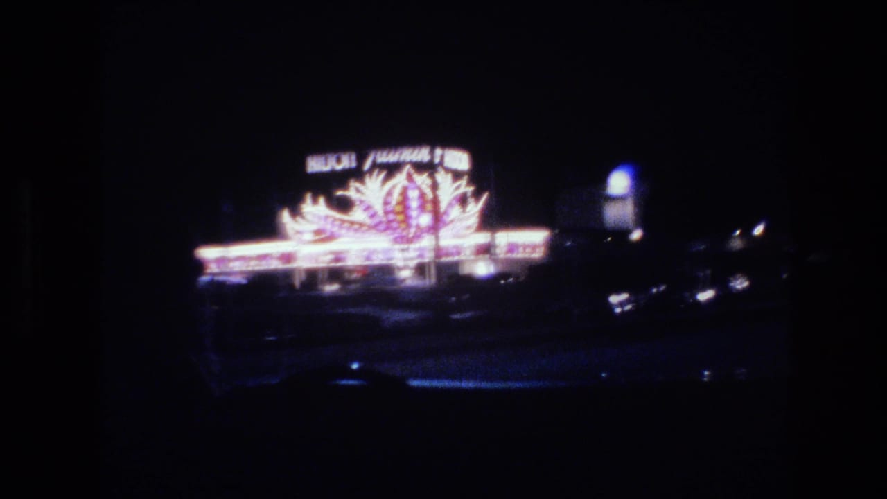 Nighttime view from inside a car of a large neon sign with stylized flame or fan shapes in pink, orange, and white against a dark sky.