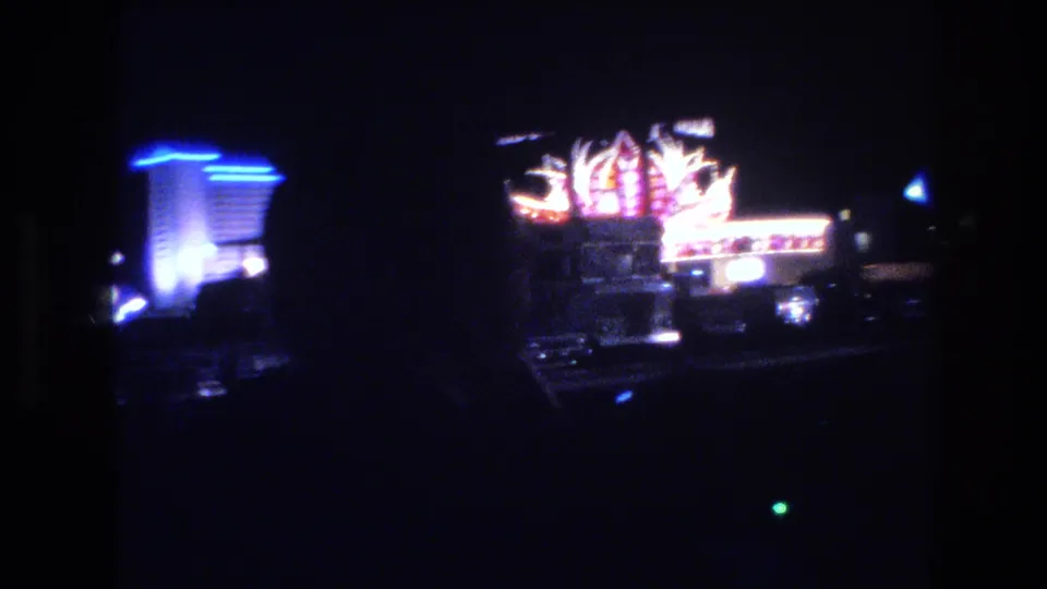 1977 Las Vegas night drive footage showing two distinct neon-lit structures: a large decorative fan-motif sign at center and a tall building with blue neon at left.