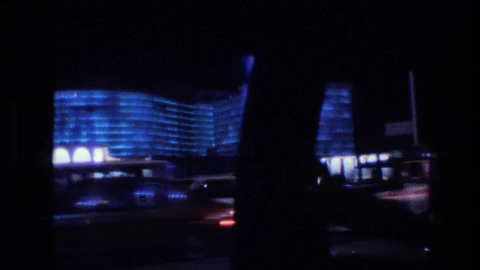 1977 Las Vegas night drive seen through a vehicle window, with a blue-lit curved hotel facade, passing traffic, and dark window framing.