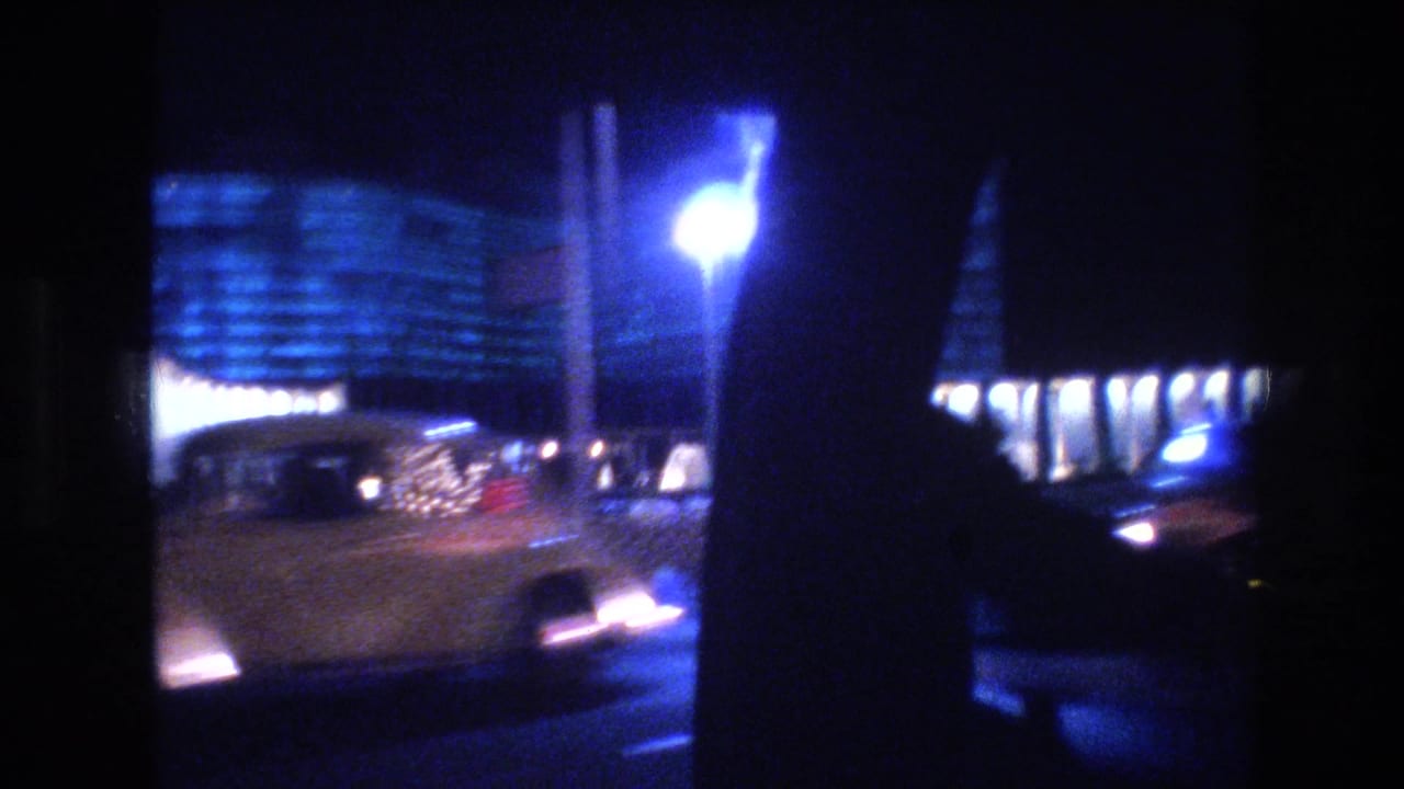 Moving 1977 Las Vegas passenger-window view with a bright streetlamp, illuminated curved facade, traffic, and heavy low-light blur.