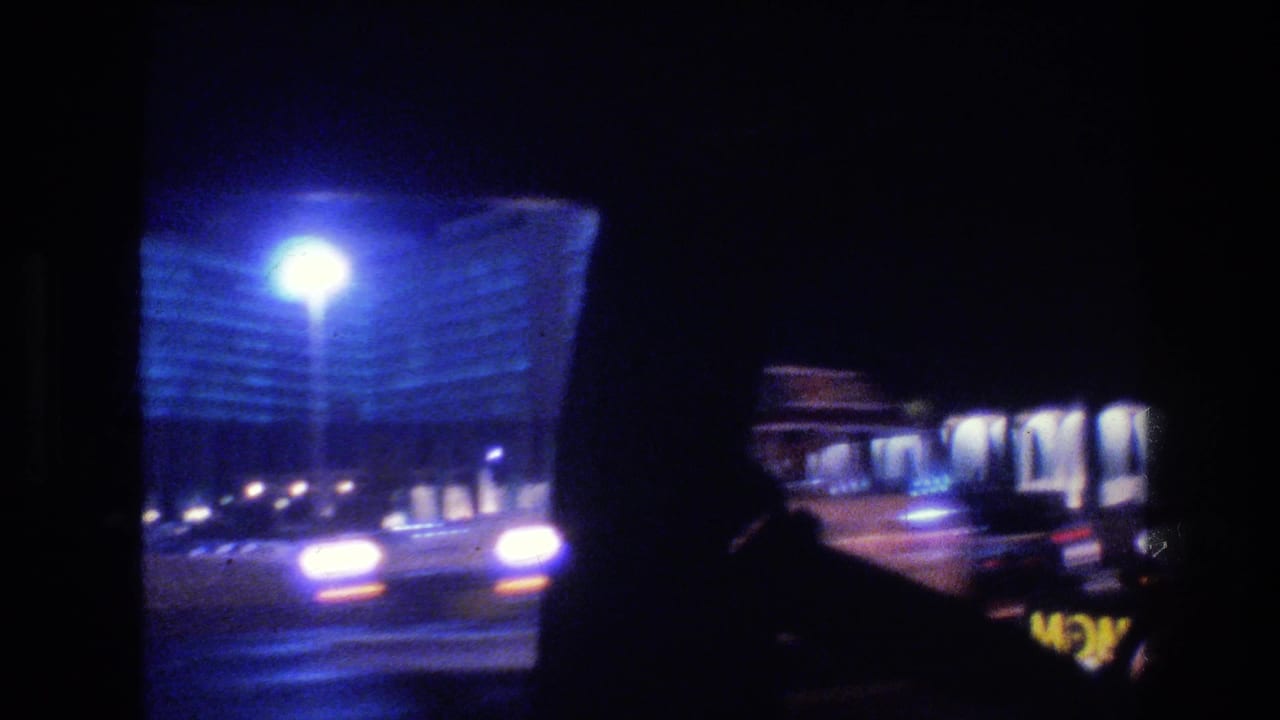 1977 Las Vegas night traffic seen from inside a car, with oncoming headlights, streetlamp flare, and a blue-lit building in the background.