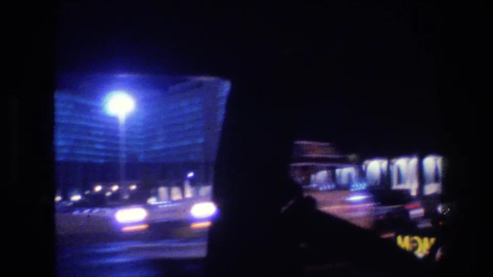 1977 Las Vegas night traffic seen from inside a car, with oncoming headlights, streetlamp flare, and a blue-lit building in the background.