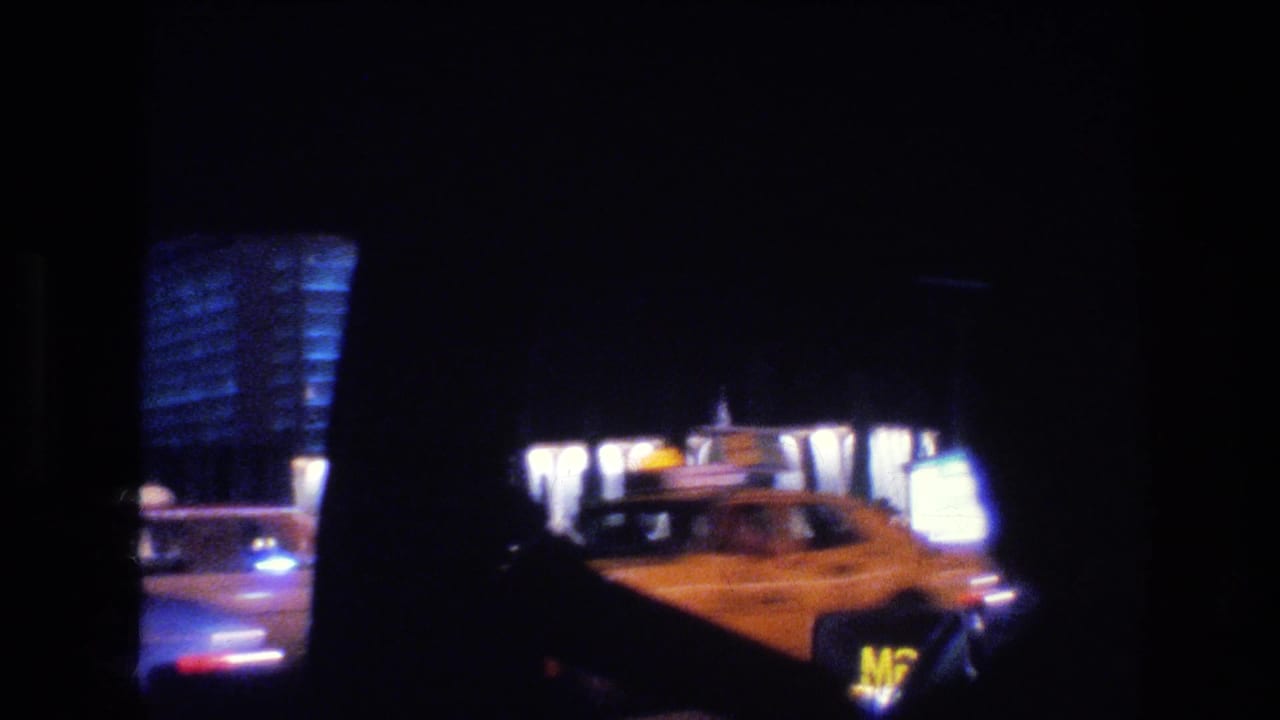 1977 Las Vegas passenger-window frame with a yellow car in the foreground, illuminated building windows, and blue-lit facade fragments at night.