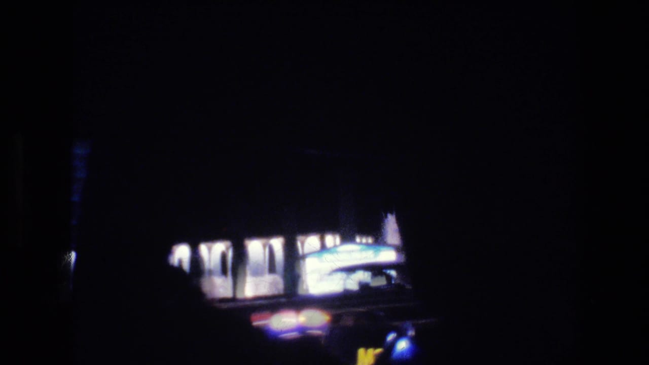 Dark street view from inside a vehicle with a lit roadside building and blurred car lights.