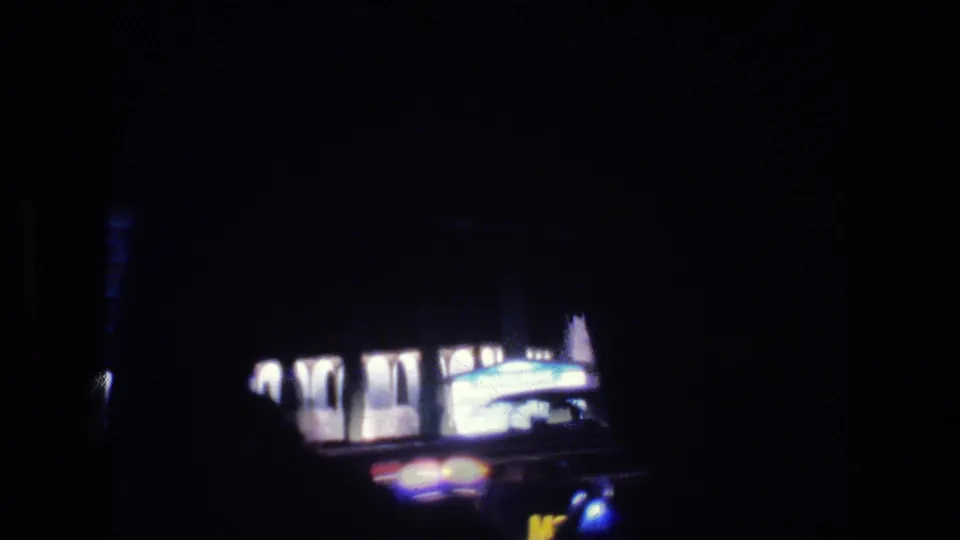 1977 Las Vegas night-drive frame showing a dim vehicle interior edge, a lit roadside facade, and blurred car lights through the window.