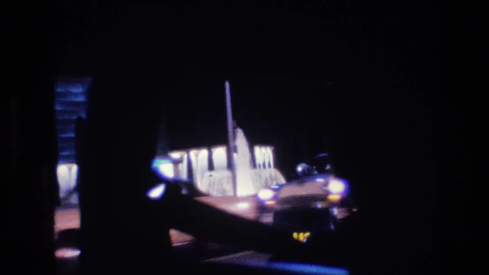 1977 Las Vegas nighttime street view with a car, glowing headlights, and a bright roadside building partly obscured by dark window edges.