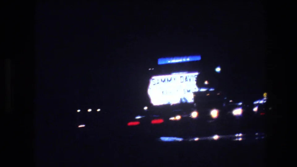1977 Las Vegas night-drive frame with a bright roadside sign, scattered car lights, and deep blue-black shadow across the street.