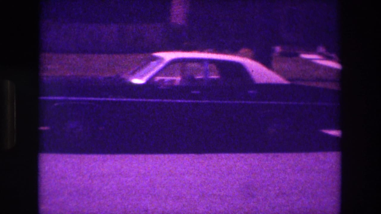 Side view of a dark sedan passing across the street beside a sidewalk and grassy area.
