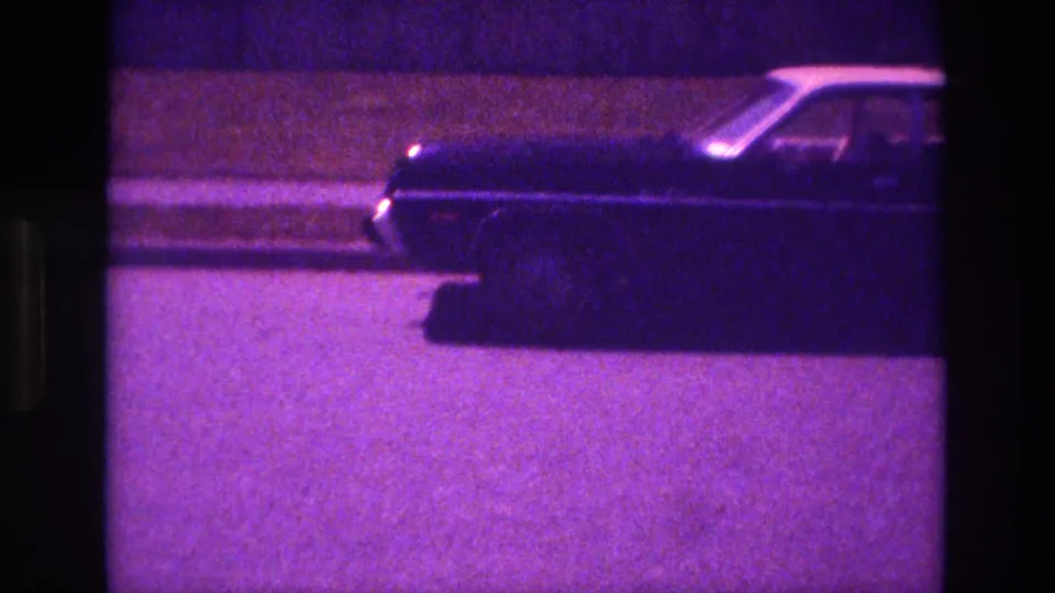 1981 San Diego 8mm frame showing the sedan continuing rightward past a sidewalk under dim ambient light.