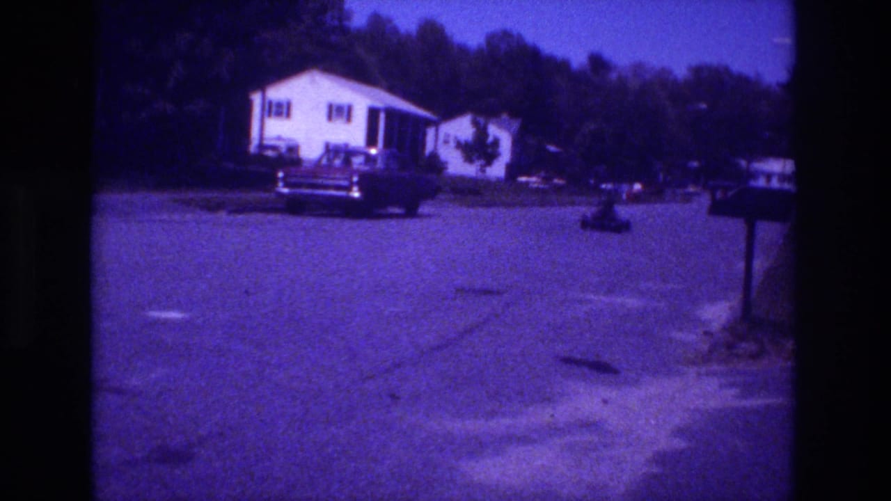 8mm home movie frame, likely San Diego USA circa 1981, showing a white single-story ranch house, a large parked American sedan, and a mailbox post on a cracked asphalt street with a dense treeline.