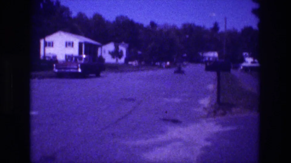 Static 8mm home movie frame from a San Diego suburban street circa 1981, showing a white ranch house, a large parked sedan, and a roadside mailbox post under a dense mature treeline.