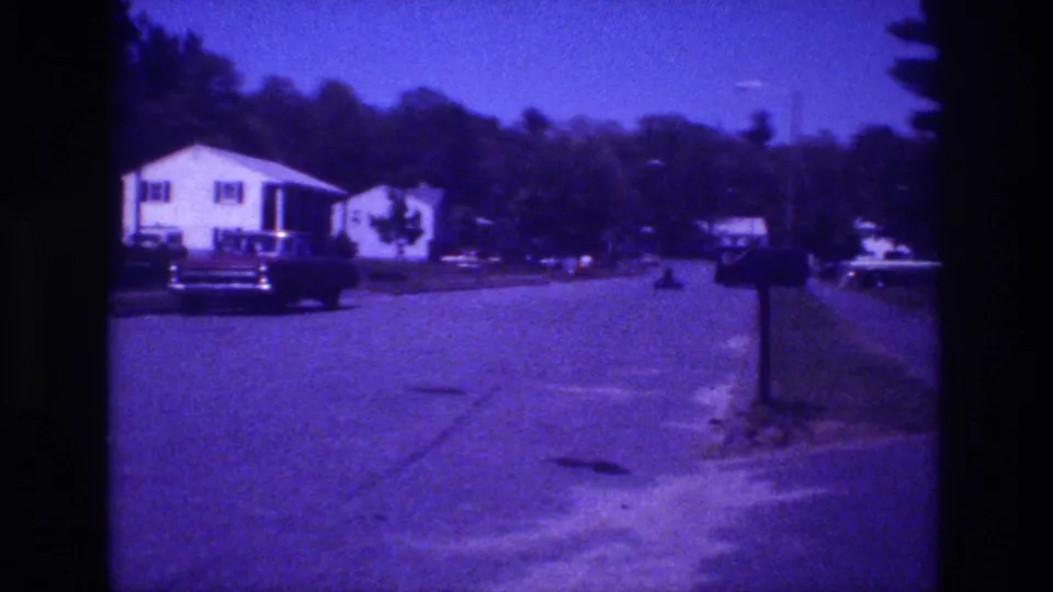 8mm home movie frame from a San Diego residential street circa 1981, showing a white single-story house, a large American car, a curbside mailbox, and additional parked vehicles visible in the background.