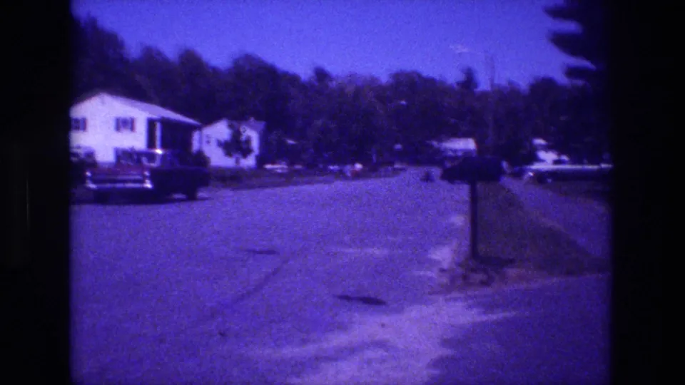Closing 8mm home movie frame from what appears to be a San Diego suburb circa 1981, showing a white ranch house, a large parked American car, and a curbside mailbox post with mature trees and blue sky.