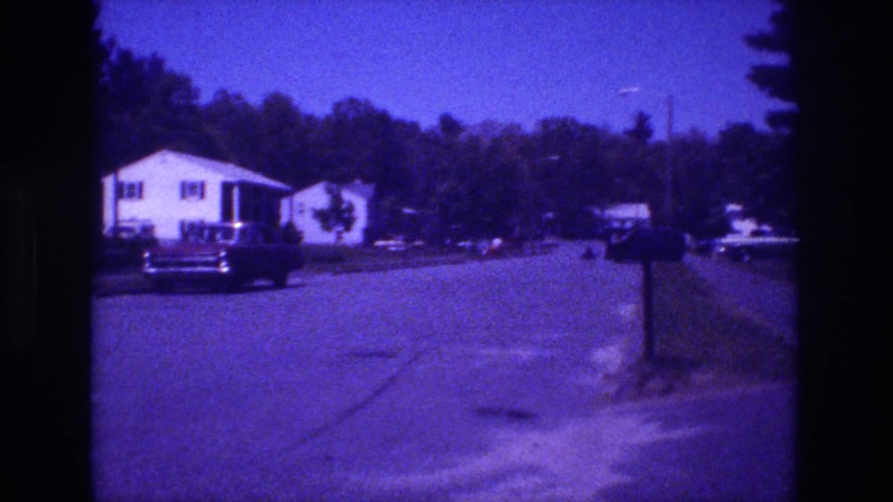1981 8mm home movie frame — white-sided suburban house, large dark sedan parked curbside, dense conifer backdrop, pronounced purple-blue film drift throughout