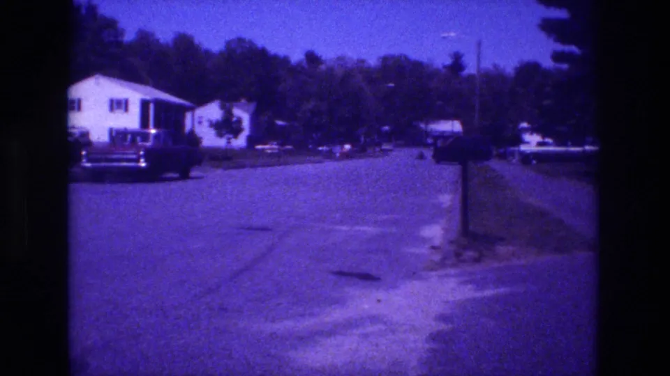 1981 8mm footage — white house with dark window accents, large dark sedan near driveway, open street foreground, conifer tree line in background, deep blue-purple color cast