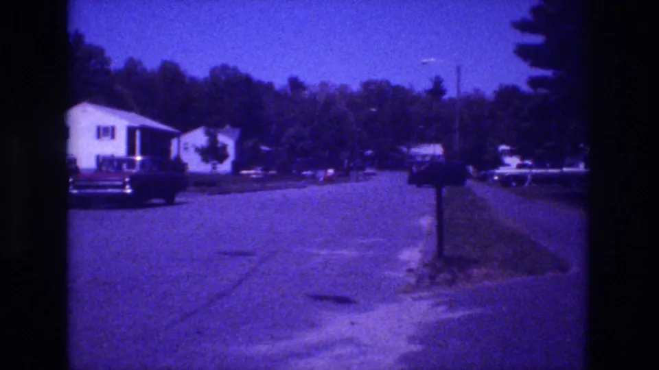 1981 8mm home movie — white-sided house, dark American sedan curbside, roadside mailbox post in mid-ground, tree-lined backdrop, heavy purple color drift throughout