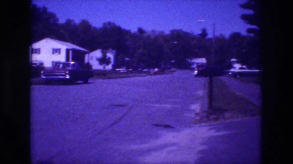 1981 8mm footage — near-identical composition to preceding frames, confirming a fully static camera; white house, dark sedan, mailbox post, tree line, purple film drift
