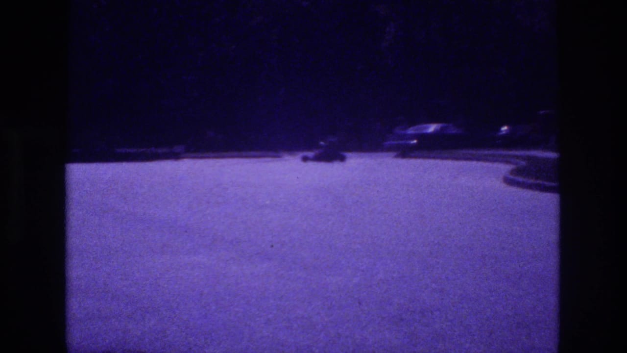 Small go-kart and parked cars on a purple-cast open parking lot at dusk, 8mm film.