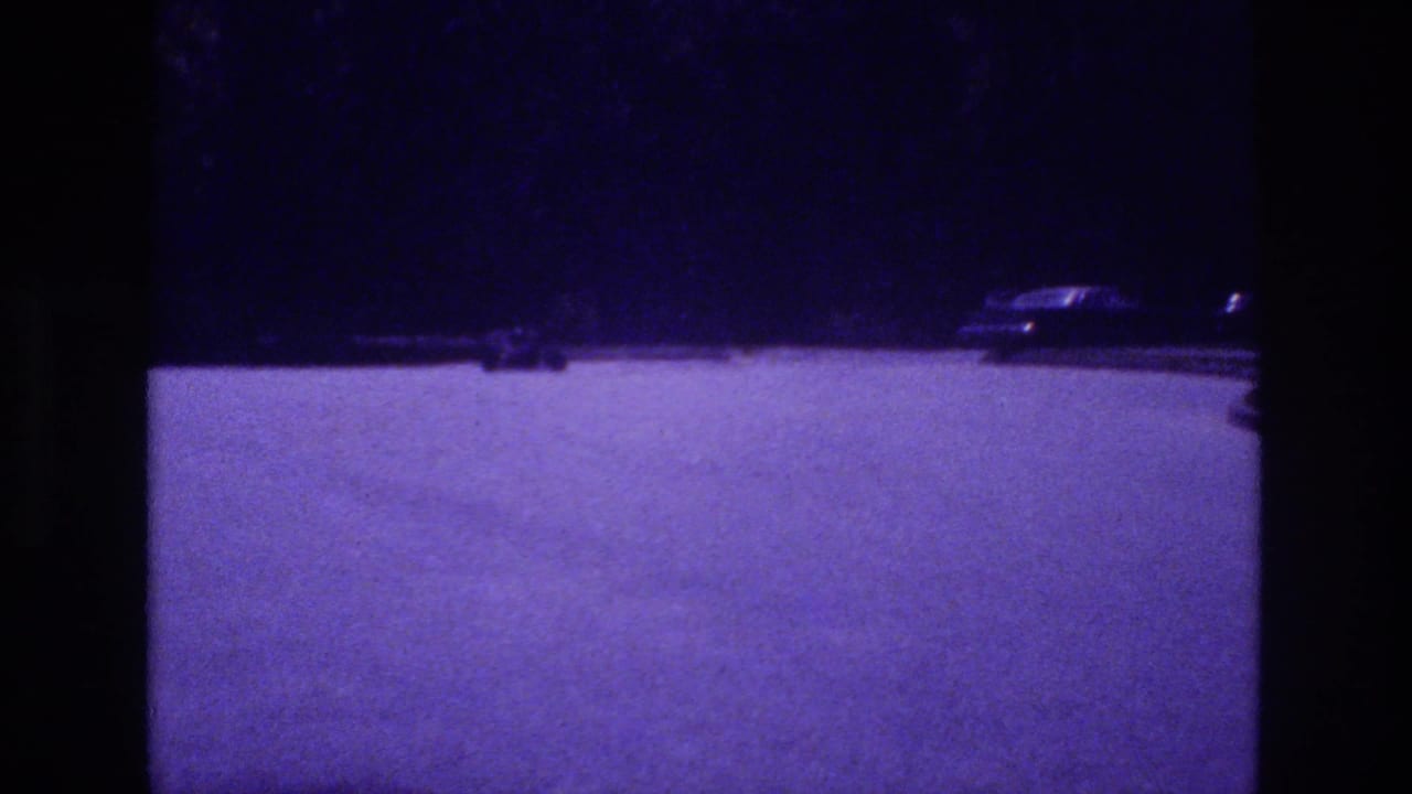 Wide purple-cast parking lot at dusk, go-kart shifted position, parked cars faintly visible upper right, 8mm film.