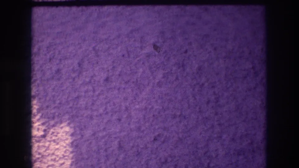 Textured popcorn stucco wall from 1982 Acapulco 8mm footage showing a bright light pool emerging at lower center against a deep purple shadow field.