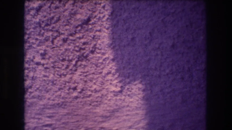 1982 Acapulco 8mm home movie frame showing a textured stucco wall with a broad warm-lit lower-left zone and deep purple shadow upper-right, separated by a diagonal shadow line.