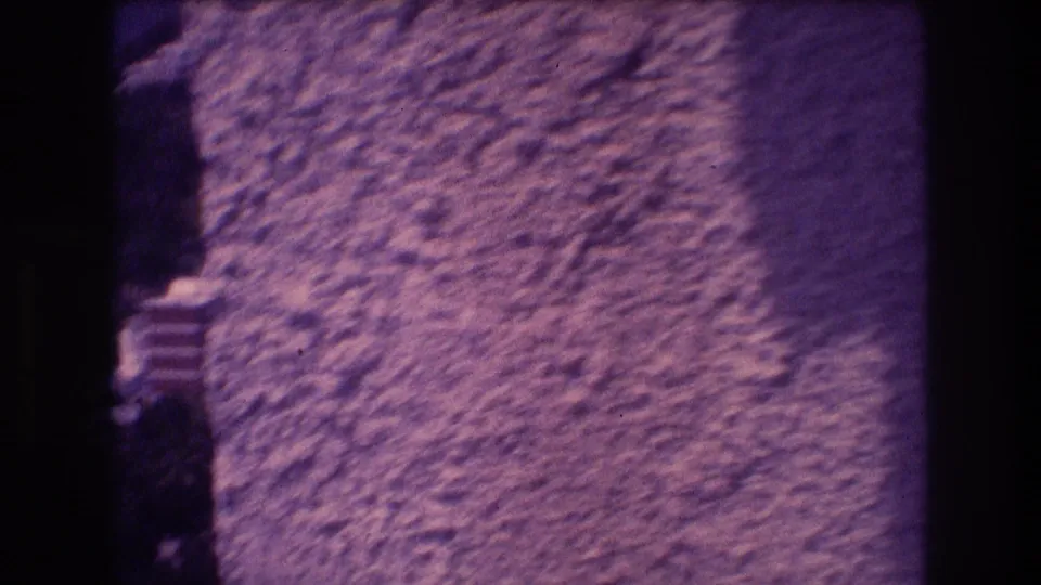 Final frame of 1982 Acapulco 8mm sequence showing a textured stucco wall, a dark vertical edge or corner at left, warm light on the right surface, and a small unidentified object at lower center.