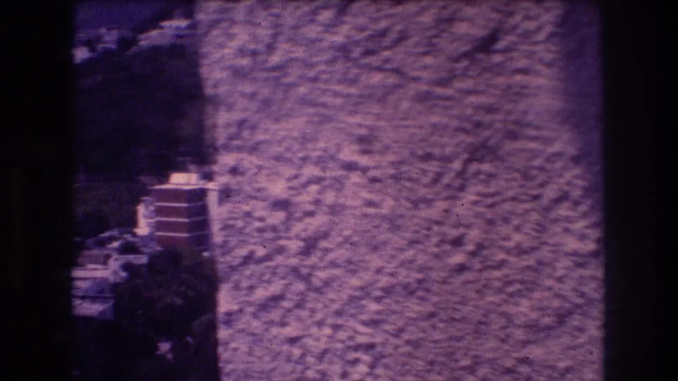 8mm home movie frame, Acapulco 1982 — textured stucco wall obstructs an opening view of terraced apartment buildings on a steep hillside.