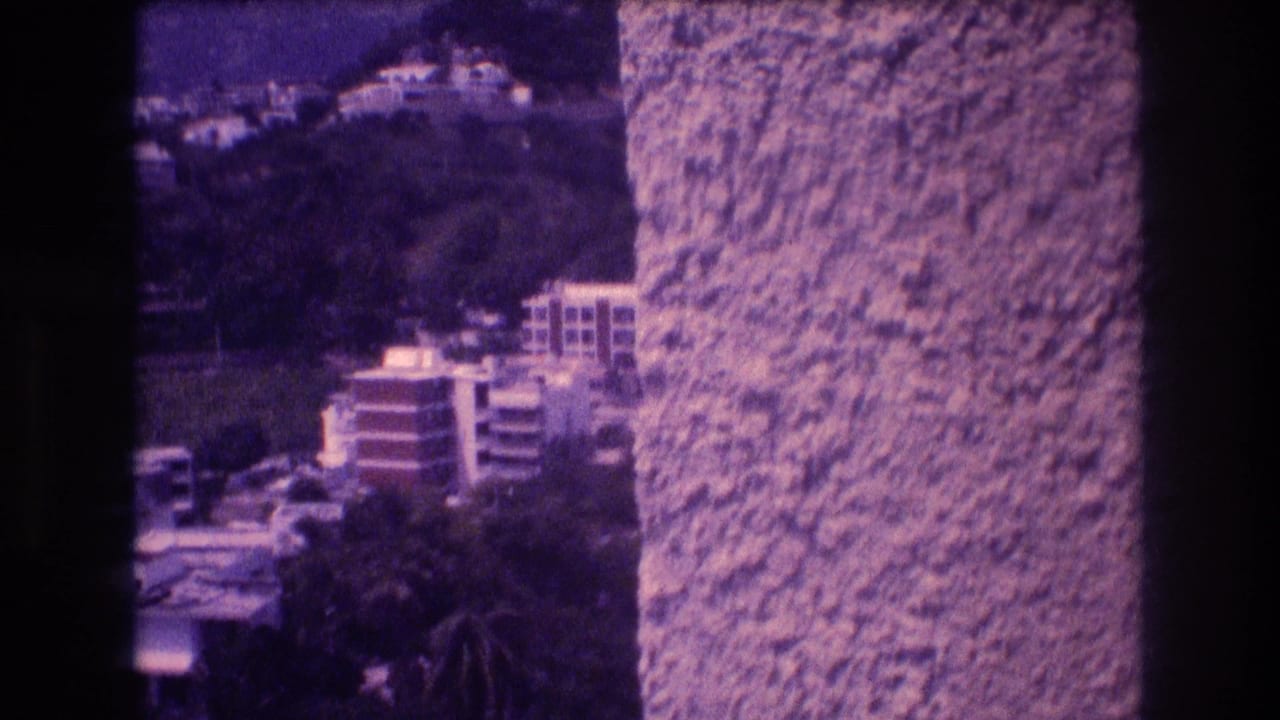 Mid-pan 8mm frame showing terraced hillside apartment buildings emerging from behind a stucco wall, Acapulco, 1982.