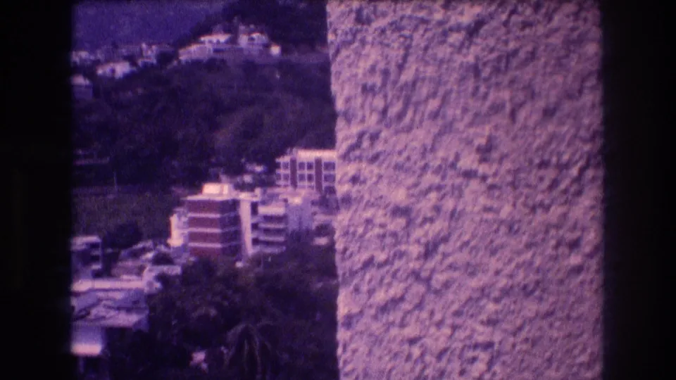 Mid-pan 8mm frame showing terraced hillside apartment buildings emerging from behind a stucco wall, Acapulco, 1982.