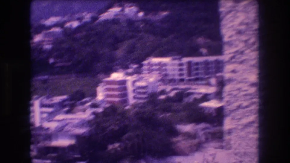 8mm pan frame showing terracotta apartment buildings terraced into a steep Acapulco hillside with tropical vegetation and mountain background, 1982.