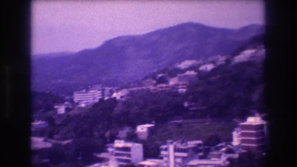 Opening frame of an 8mm home movie pan across Acapulco, Mexico, 1982 — white multi-story buildings on a green hillside, heavy purple dye shift and edge vignetting.