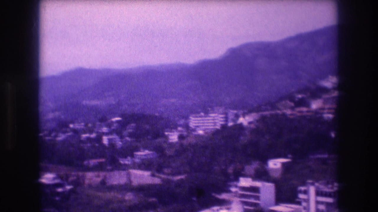 Second pan frame across Acapulco hillside, 1982 — clustered white buildings set against green mountains, characteristic 8mm purple emulsion aging and soft focus.