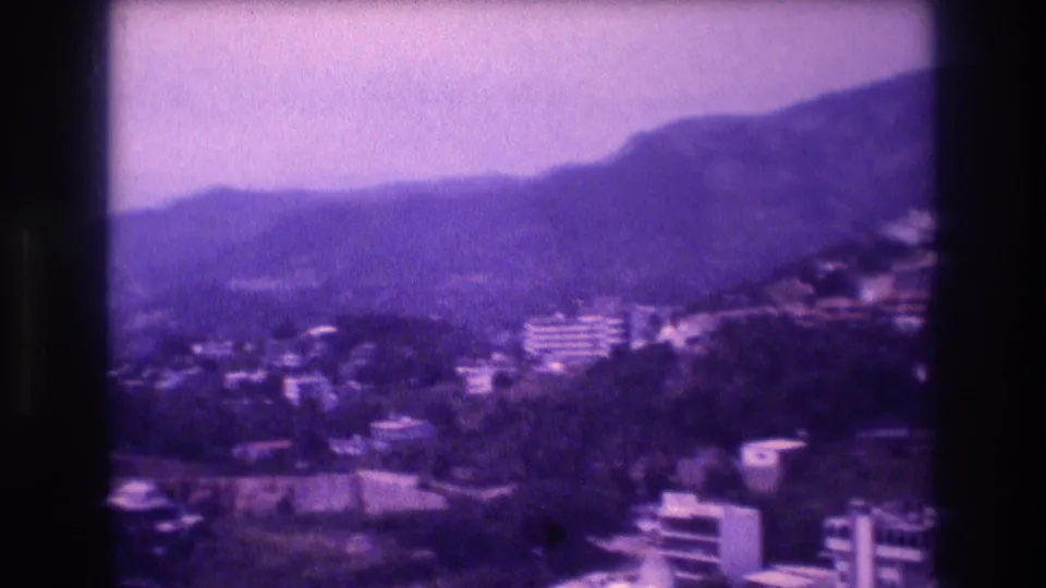 Second pan frame across Acapulco hillside, 1982 — clustered white buildings set against green mountains, characteristic 8mm purple emulsion aging and soft focus.