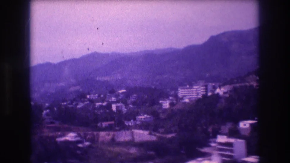 Wider pan frame of Acapulco, Mexico, 1982 — city buildings spread across lower hillside slopes, tall mountain peaks behind, strong purple dye shift and visible film grain.