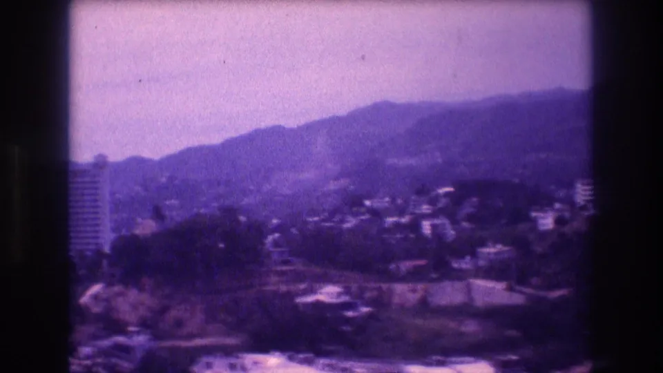 Final pan frame of Acapulco hillside, 1982 — a tall high-rise tower rises on the right among lower structures, against a mountain backdrop with purple 8mm emulsion aging.
