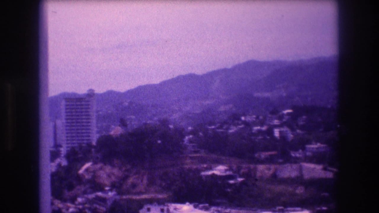 8mm home movie frame from what appears to be Acapulco, Mexico, circa 1982, showing a tall hotel tower against forested hills under a hazy sky, with a strong purple color cast.