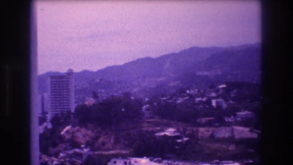 8mm home movie frame from what appears to be Acapulco, Mexico, circa 1982, showing a tall hotel tower against forested hills under a hazy sky, with a strong purple color cast.