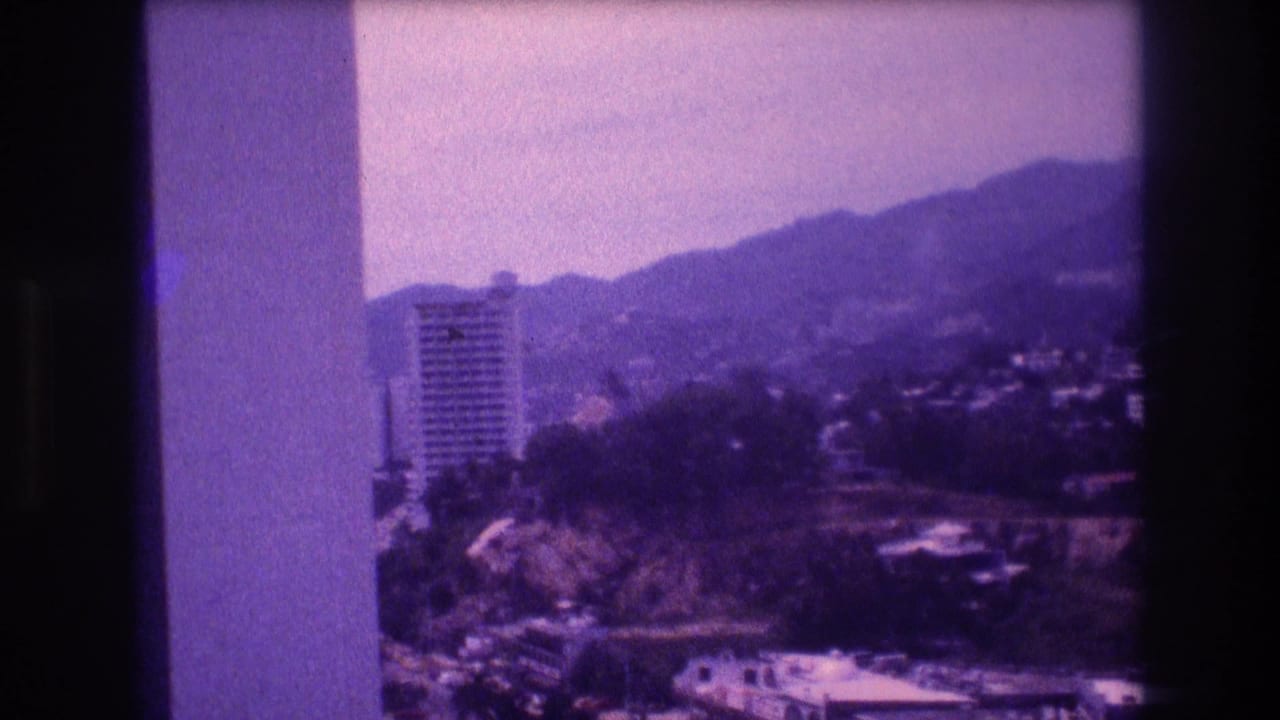 8mm home movie frame circa 1982 showing a high-rise tower set against rolling hills in what appears to be Acapulco, with a concrete balcony structural element entering from frame left.