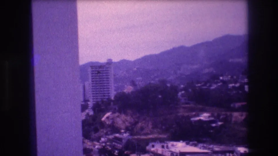 8mm home movie frame circa 1982 showing a high-rise tower set against rolling hills in what appears to be Acapulco, with a concrete balcony structural element entering from frame left.