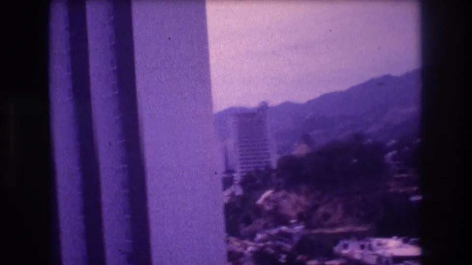 8mm home movie frame circa 1982 mid-pan showing the same hotel tower now positioned toward frame left, forested hills filling the background, in what appears to be Acapulco.