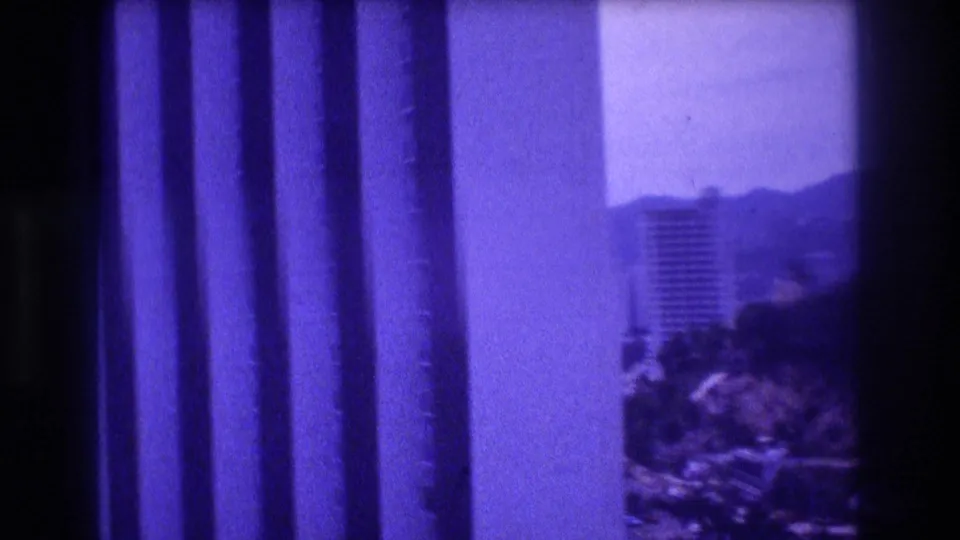 8mm home movie end-frame circa 1982 showing foreground vertical window or balcony structural elements in purple-blue tones, with the hotel tower and hillside glimpsed at right through the gap in what appears to be Acapulco.