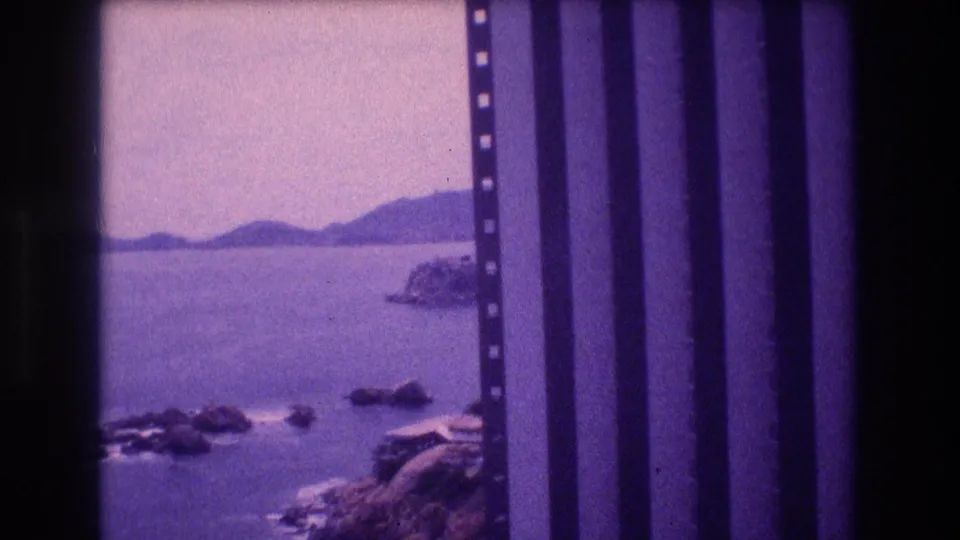 8mm home movie frame from Acapulco, 1982; elevated lookout view shows rocky shoreline, calm bay, and distant hills under a pale sky.