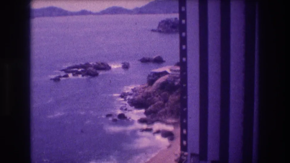 High-vantage 8mm home movie frame from Acapulco, Mexico, c. 1982, showing rocky offshore outcrops, a sandy beach, a distant headland, and purple dye-shifted film.
