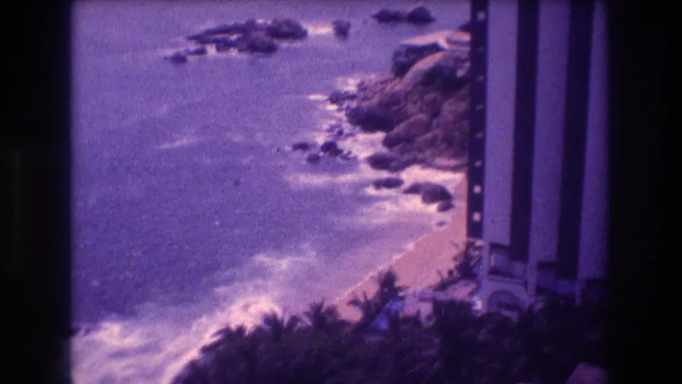Elevated 8mm home movie shot from Acapulco, Mexico, c. 1982, showing white surf on a rocky shore, palm trees, and a multi-story resort building at the right margin.