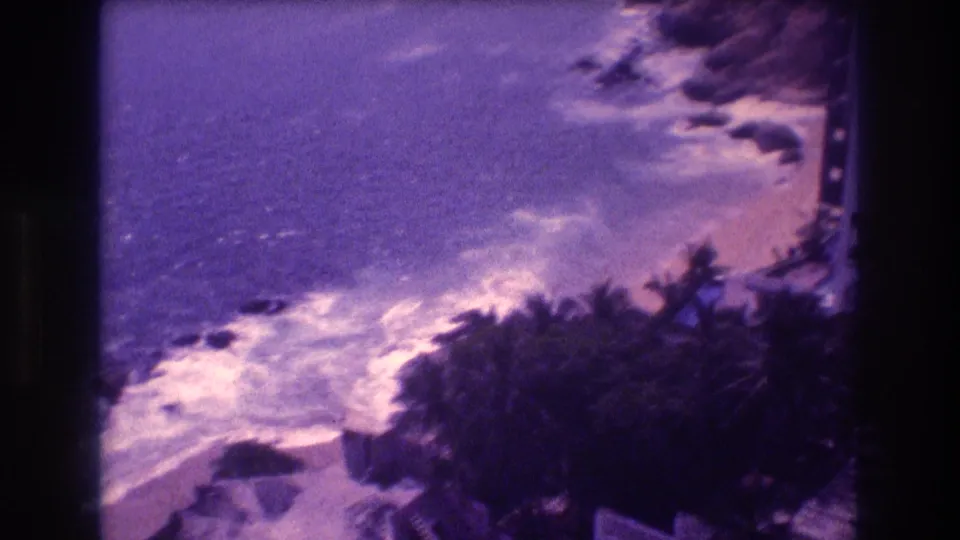8mm home movie frame from Acapulco, c. 1982, angled down over palm trees toward a rocky shoreline with breaking surf and a narrow sandy beach, with strong purple dye-shift.