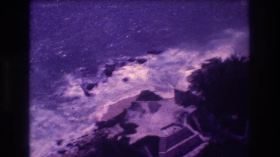 Terminal frame of an 8mm Acapulco home movie, c. 1982, showing rough surf on a rocky shore, dense palm vegetation, and a low-rise resort structure at lower right.