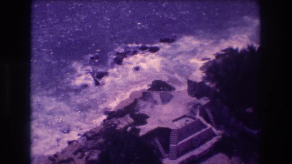 1982 8mm home movie frame showing breaking surf on rocky Acapulco coast from a bridge overlook, with stepped structure and tropical vegetation below.